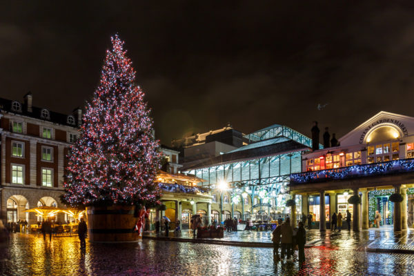 An image of the entrance to Covent garden market at night time. There is a christmas tree with lights on and a few groups of people