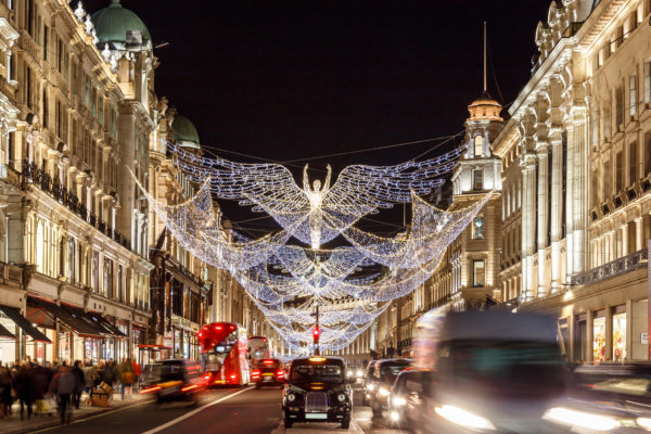 Christmas lights in Mayfair. An angel and its wings hanging over the road between the shops on either side. There is moving traffic at the bottom with a red bus and black cab